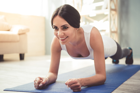 Beautiful young woman in sportswear is smiling while doing plank on exercise mat at home