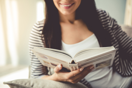 Cropped image of beautiful young woman in casual clothes reading a book and smiling while sitting on couch at homeの写真素材