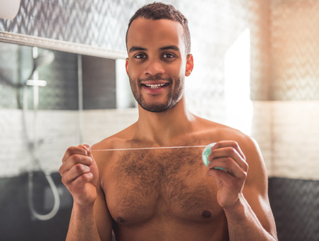 Handsome Afro American man is holding a dental floss, looking at camera and smiling while standing in bathroomの写真素材