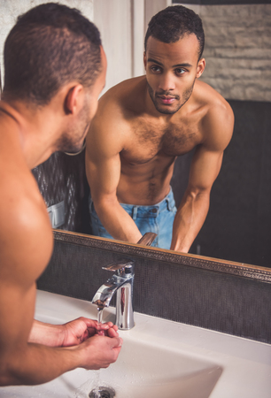 Handsome Afro American man is washing himself and looking into the mirror in bathroomの写真素材