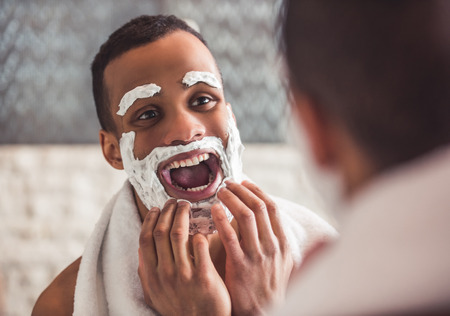 Handsome Afro American man is making faces and having fun using shaving foam while looking into the mirror in bathroomの写真素材