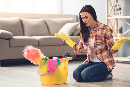 Beautiful tired young woman in protective gloves is sitting confused near the bucket with things for cleaning while cleaning her houseの写真素材