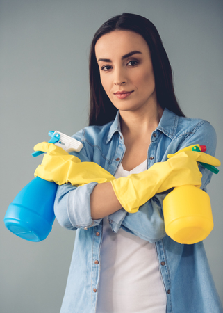 Beautiful young woman in protective gloves is holding a detergents and looking at camera, on gray backgroundの写真素材