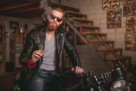 Handsome bearded man in leather jacket and sun glasses is smoking a cigarette while sitting on the motorcycle in the repair shopの写真素材