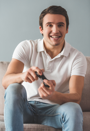 Handsome young man is playing game console and smiling while sitting on couch at homeの写真素材