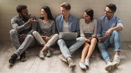 Group of beautiful students in casual clothes using gadgets, talking and smiling while sitting together on the floorの写真素材