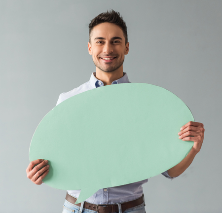 Attractive young man is holding a speech bubble, looking at it and smiling, on gray background