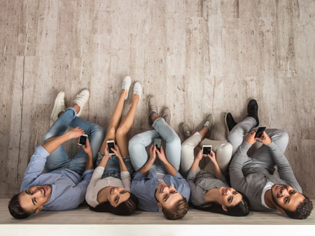 Top view of beautiful young people in casual clothes using smartphones, looking at camera and smiling while sitting together on the floorの写真素材