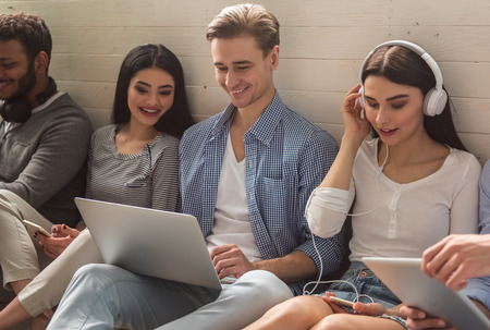 Group of beautiful students in casual clothes using gadgets and smiling while sitting together on...