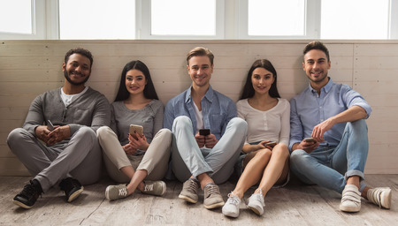Group of beautiful young people in casual clothes using smartphones, looking at camera and smiling while sitting together on the floorの写真素材
