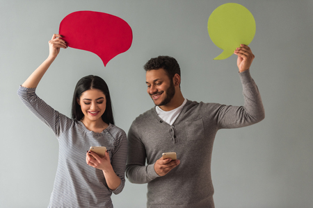 Beautiful Afro American couple is holding speech bubbles, using smartphones and smiling, on gray backgroundの写真素材