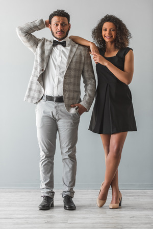 Full length portrait of beautiful Afro American couple in formal wear looking at camera, on gray background. Guy is embarrassed, girl is smilingの写真素材