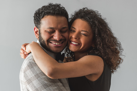 Beautiful Afro American couple in formal wear is hugging with closed eyes and smiling, on gray backgroundの写真素材