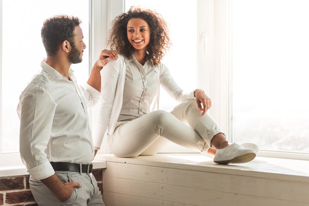 Beautiful Afro American couple in smart casual wear is talking and smiling while sitting near the windowの写真素材