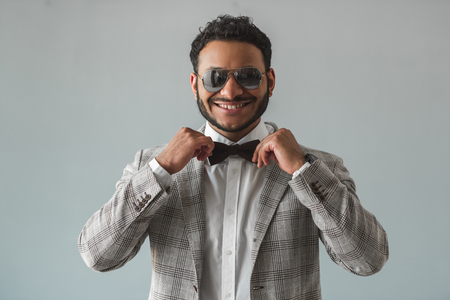 Stylish Afro American guy in suit and sun glasses is adjusting his bow tie and smiling, on gray backgroundの写真素材