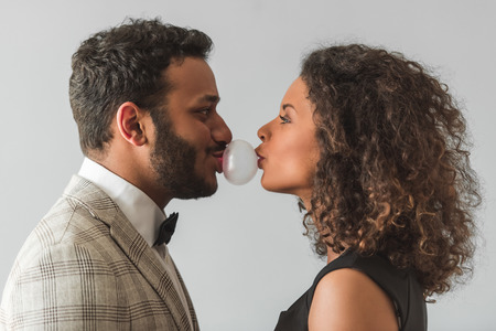 Side view of beautiful Afro American couple in formal wear chewing a bubble gum and making a bubble,...