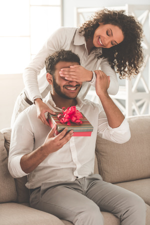 Happy Afro American couple at home. Beautiful girl is giving a gift box to her boyfriendの写真素材