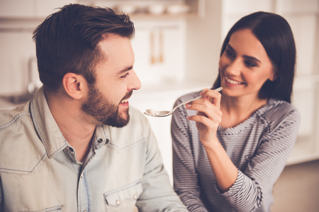 Beautiful couple is eating and smiling while sitting in kitchen at home. Girl is feeding her husbandの写真素材