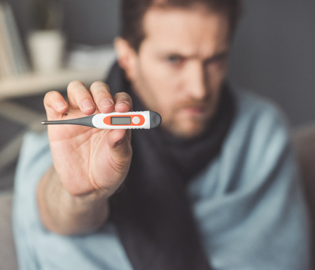 Handsome sick man is holding a thermometer while sitting on couch covered in blanket. Thermometer in...