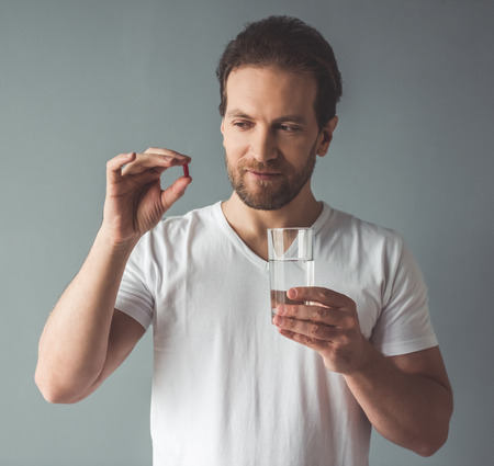 Handsome man is holding a pill and a glass of water, on gray backgroundの写真素材