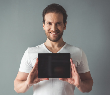 Handsome man is holding a digital tablet, looking at camera and smiling, on gray backgroundの写真素材