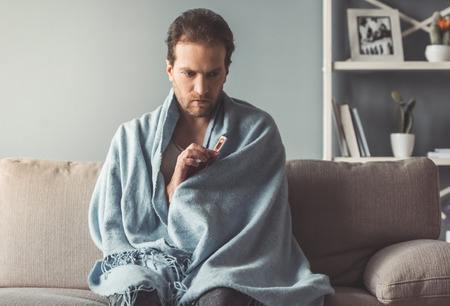 Handsome sick man is holding a thermometer while sitting on couch covered in plaidの写真素材