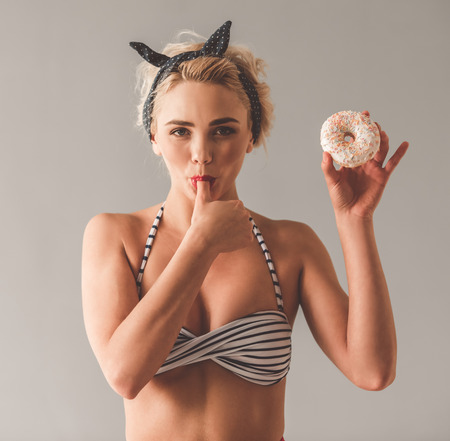 Stylish young girl in summer clothes is holding doughnut, licking her finger and smiling, on gray backgroundの写真素材