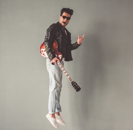 Handsome young man in leather jacket and sun glasses is holding a guitar and showing rock sign while...
