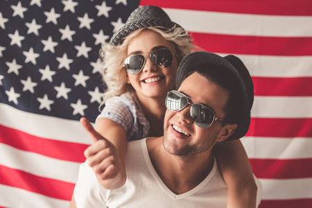 Stylish young couple in hats and sun glasses is hugging, looking at camera and smiling, on American flag backgroundの写真素材