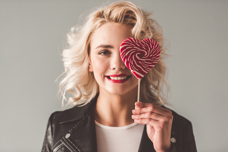 Stylish young girl in leather jacket is holding a heart-shaped lollipop and smiling, on gray backgroundの写真素材