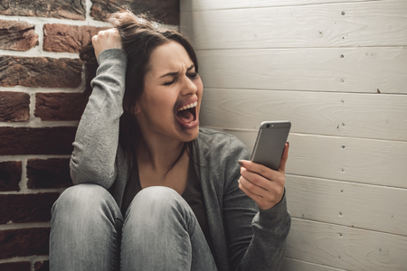 Girl being struck. Portrait of young girl with bruises on her face holding a mobile phone and screaming while sitting on the floorの写真素材