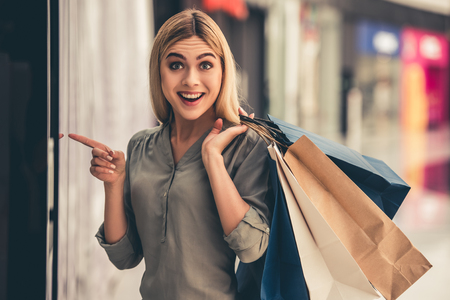 Attractive girl is holding shopping bags, pointing, looking at camera and smiling while doing shopping in the mallの写真素材