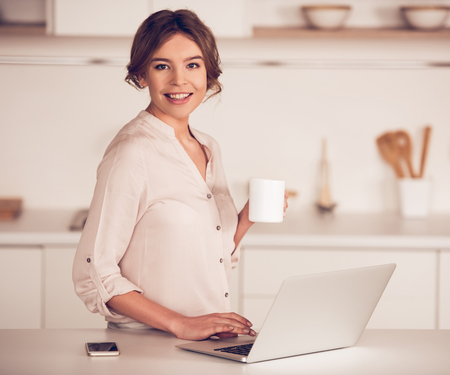Beautiful business woman in casual clothes is holding a cup, looking at camera and smiling while working with a laptop in kitchenの写真素材