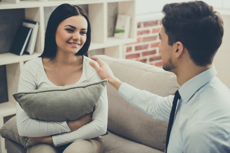 Beautiful young woman is hugging a cushion and smiling while sitting on the couch at the psychologistの写真素材