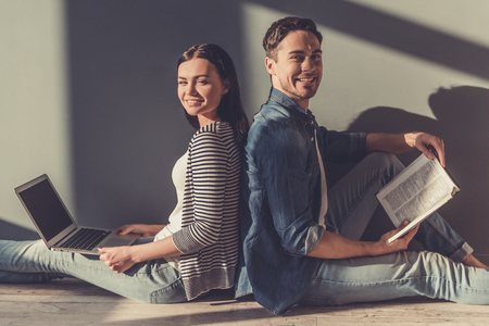 Beautiful young couple is looking at camera and smiling while sitting on the floor back to back. Girl is using laptop, guy is reading bookの写真素材