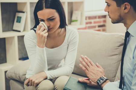 Beautiful young woman is crying while sitting on the couch at the psychologistの写真素材