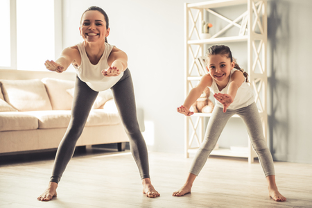 Beautiful young woman and charming little daughter are smiling while doing yoga together at homeの写真素材