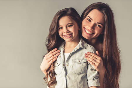 Charming little girl and her beautiful young mom are hugging, looking at camera and smiling, on gray backgroundの写真素材