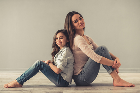 Charming little girl and her beautiful young mom are looking at camera and smiling while sitting back to back on the floorの写真素材