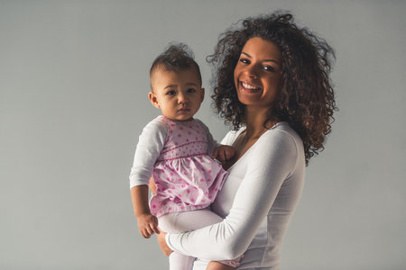 Beautiful young Afro American woman and her cute baby are looking at camera and smiling, on gray backgroundの写真素材
