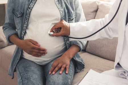 Handsome Afro American doctor in white coat is consulting beautiful pregnant woman in his officeの写真素材