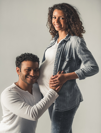 Handsome Afro American man is listening to his beautiful pregnant wife's tummy, feeling pleasedの写真素材