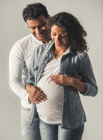 Handsome Afro American man and his beautiful pregnant wife are hugging and smiling, on gray backgroundの写真素材