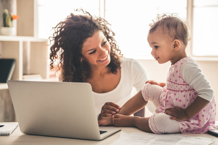 Beautiful young Afro American woman and her cute baby are using a laptop and smiling while spending time together at homeの写真素材