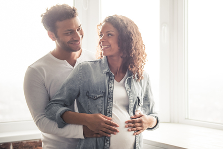 Handsome Afro American man and his beautiful pregnant wife are hugging and smiling while standing near the window at homeの写真素材