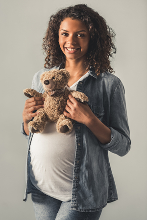 Beautiful pregnant Afro American woman is holding a teddy bear, looking at camera and smiling, on gray backgroundの写真素材