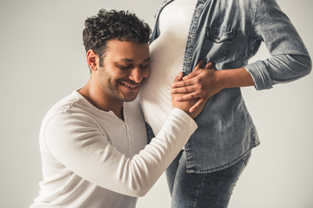 Handsome Afro American man is listening to his beautiful pregnant wife's tummy, feeling pleasedの写真素材