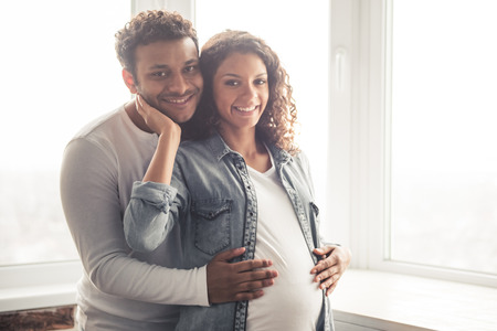 Handsome Afro American man and his beautiful pregnant wife are hugging and smiling while standing near the window at homeの写真素材