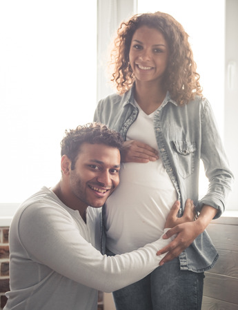 Handsome Afro American man and his beautiful pregnant wife are hugging and smiling while standing near the window at homeの写真素材