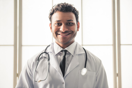 Handsome Afro American doctor in white coat is looking at camera and smiling while standing in officeの写真素材